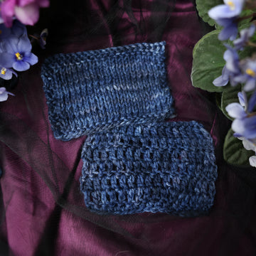 A dark blue crochet and knit swatches that has hues of dark grey. This skein is on top of a dark purple tablecloth and dark blackish blue tablecloth with blue and purplish pink flowers laying next to the skein on both sides.