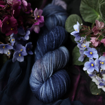 A dark blue Worsted yarn that has hues of grey. This skein is on top of a dark purple tablecloth and dark blackish blue tablecloth with blue and purplish pink flowers laying next to the skein on both sides.