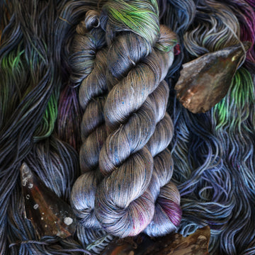 Silk yarn colored with different shades of grey and streaks of green, and dark pink, also there are also blue and purple speckles. These skeins are on top of unwound skeins of the same color way with some shells on top.