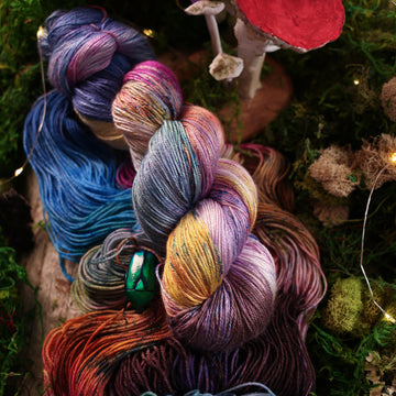 Silk yarn colored with a hot pink, light blue, dark blue/purple, with some green and orange/brown colors, on top of the same yarn but unwound, with moss underneath and some fairy lights around the moss and a small glass looking beetle and a mushroom in the back.