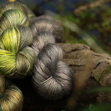 A dark grey Silk yarn with hints of green colors, on top of a tree limb that is hovering over a pond, with different plants around the pond, and around the yarn.