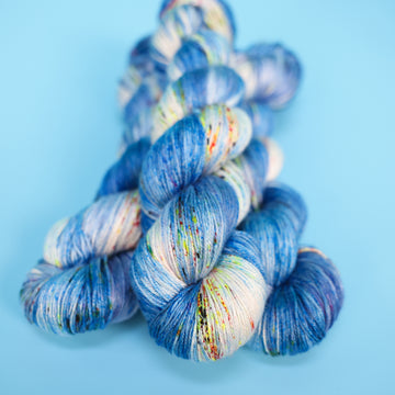 Silk yarn colored with different shades of blue and white with multicolored speckles on the portion of the yarn that Is white. all on top of a light blue background.