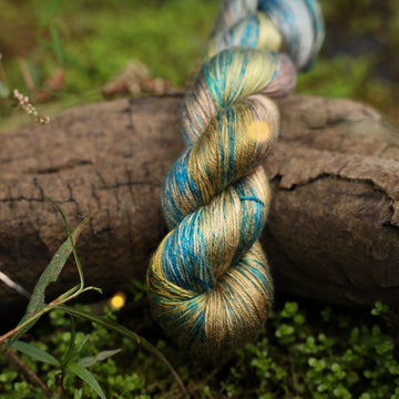 A Silk yarn skein colored with green, yellow and grey colors, with blue speckles, on top of a tree limb that is hovering over a pond, with different mossy plants around the pond, and around the yarn.