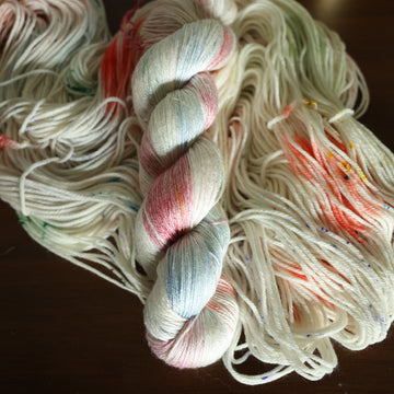 Silk white yarn with mixed colors of light pink purple and red/orange with speckles, on top of an unwound version of that same yarn. all on top of a dark wooden table with a couple of leaves of a plant overhead in a gold pot.