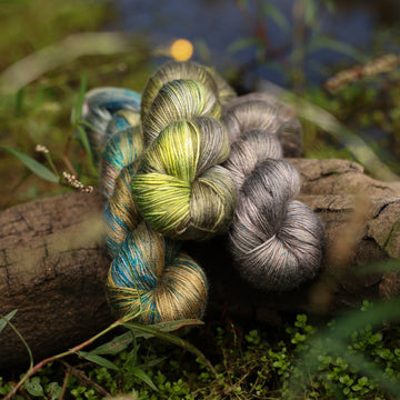 Different Silk yarn skeins, one colored a lime green and grey, one colored a dark grey, and one colored green with blue speckles, on top of a tree limb that is hovering over a pond, with different mossy plants around the pond, and around the yarn.