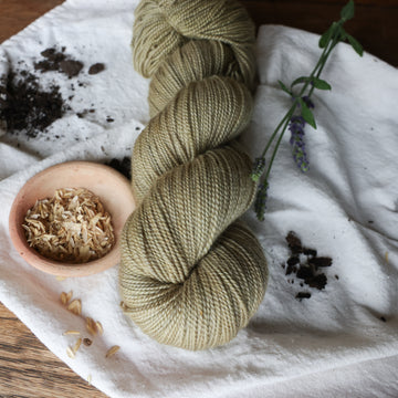 A very calm pretty light brownish green colored fingering yarn skein on top of a white tablecloth with some flowers and rice grains and soil in the corner on top of a wooden table.