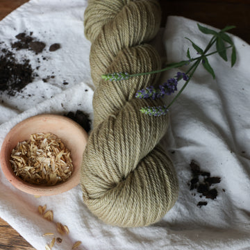 A very calm pretty light brownish green colored worsted yarn skein on top of a white tablecloth with some small purple flowers and rice grains and soil in the corner on top of a wooden table.