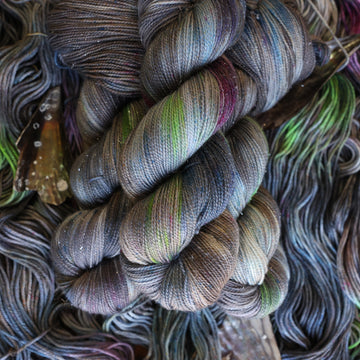 Sparkle yarn colored with different shades of grey and streaks of green, and dark pink, also there are also blue and purple speckles. These skeins are on top of unwound skeins of the same color way with some shells on top.
