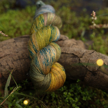A Sparkle yarn skein colored with green, yellow and grey colors, with blue speckles, on top of a tree limb that is hovering over a pond, with different mossy plants around the pond, and around the yarn.