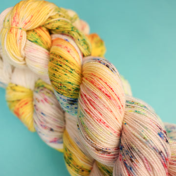 A light cream colored Sparkle yarn with different colored speckles all over and banana colored yellow at the top of the skein on top of a light blue background.