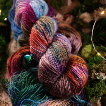 Sparkle yarn colored with a hot pink, light blue, dark blue/purple, with some green and orange/brown colors, on top of the same yarn but unwound, with moss underneath and some fairy lights around the moss and a small glass looking beetle.