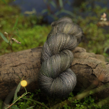 A dark grey sparkle yarn with hints of green colors, on top of a tree limb that is hovering over a pond, with different plants around the pond, and around the yarn.