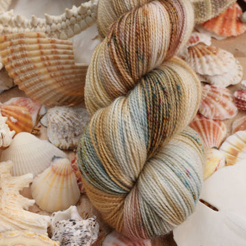 Sparkle yarn that is colored with different light browns and creme colors along with some blues and speckles of blue brown and pink. This yarn is on top of a bunch of different types  of light colored shells and sea stars and sand dollars.
