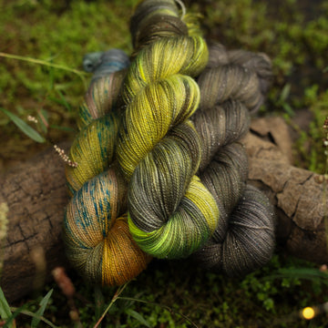 Different sparkle yarn skeins, one colored a lime green and grey, one colored a dark grey, and one colored green with blue speckles, on top of a tree limb that is hovering over a pond, with different mossy plants around the pond, and around the yarn.
