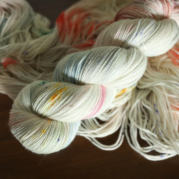 sparkle white yarn with mixed colors of light pink purple and red/orange with speckles, on top of an unwound version of that same yarn. all on top of a dark wooden table.