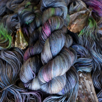 Sport yarn colored with different shades of grey and streaks of green, and dark pink, also there are also blue and purple speckles. These skeins are on top of unwound skeins of the same color way with some shells on top.