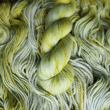 Sport yarn colored with different shades of light green and white with some dark green speckles on top of the same unwound yarn.