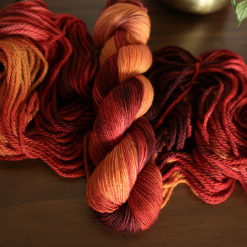 Sport yarn with red orange and yellow colors, on top of an unwound version of that same yarn. all on top of a dark wooden table with a couple of leaves of a plant overhead in a gold pot.
