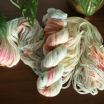 Sport white yarn with mixed colors of light pink purple and red/orange and brown with speckles, on top of an unwound version of that same yarn. all on top of a dark wooden table with a couple of leaves of a plant overhead in a gold pot.