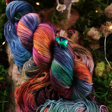 Sport yarn colored with a hot pink, light blue, dark blue/purple, with some green and orange/brown colors, on top of the same yarn but unwound, with moss underneath and some fairy lights around the moss and a small glass looking beetle.