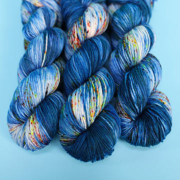 Sport yarn colored with different shades of blue and white with multicolored speckles on the portion of the yarn that Is white. all on top of a light blue background.