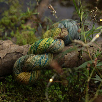 A Sport yarn skein colored with green, yellow and grey colors, with blue speckles, on top of a tree limb that is hovering over a pond, with different mossy plants around the pond, and around the yarn.