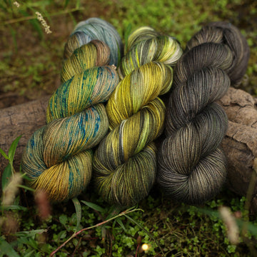 Different Sport yarn skeins, one colored a lime green and grey, one colored a dark grey, and one colored green with blue speckles, on top of a tree limb that is hovering over a pond, with different mossy plants around the pond, and around the yarn.