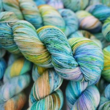 Sport yarn with light shades of blue and speckles of purple, with some light greens and browns. There are more skeins like this of different weights underneath.