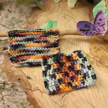A mixture of colors like burnt orange, yellow, lime green, black/grey, and teal colored knit and crochet swatches, on top of a plant with bare branches and some leaves in the corner. along with painted butterflies and leaves made of book pages, all on top of a wooden table.
