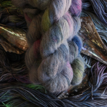 Suri yarn colored with different shades of grey and streaks of green, and dark pink, also there are also blue and purple speckles. These skeins are on top of unwound skeins of the same color way with some shells on top.