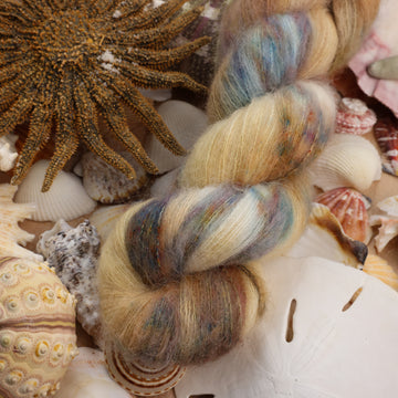 Suri yarn that is colored with different light browns and creme colors along with some blues and speckles of blue brown and pink. This yarn is on top of a bunch of different types  of light colored shells and sea stars and sand dollars.