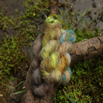 Different Suri Lace yarn skeins, one colored a lime green and grey, one colored a dark grey, and one colored green with blue speckles, on top of a tree limb that is hovering over a pond, with different mossy plants around the pond, and around the yarn.