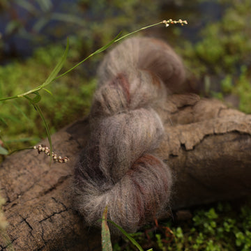 A dark grey suri lace yarn with hints of green  and brown colors, on top of a tree limb that is hovering over a pond, with different plants around the pond, and around the yarn.
