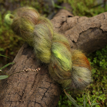 A Suri Lace yarn skein colored with different shades of green and grey, on top of a tree limb that is hovering over a pond, with different mossy plants around the pond, and around the yarn.