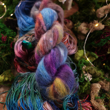 Suri yarn colored with a hot pink, light blue, dark blue/purple, with some green and orange/brown colors, on top of the same yarn but unwound, with moss underneath and some fairy lights around the moss and a small glass looking beetle.