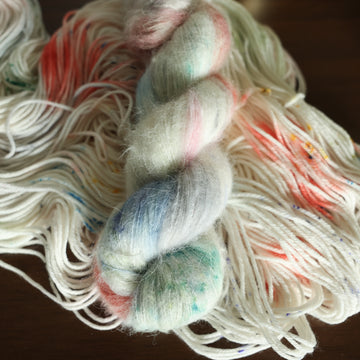 Suri white yarn with mixed colors of light pink purple and red/orange with speckles, on top of an unwound version of that same yarn. all on top of a dark wooden table.