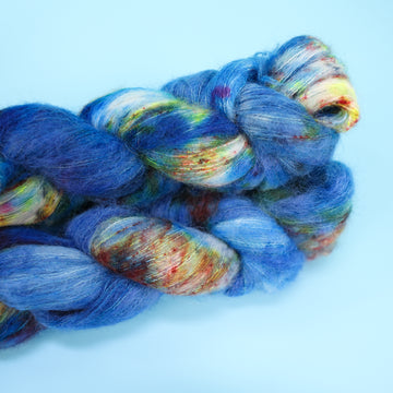 Suri yarn colored with different shades of blue and white with multicolored speckles on the portion of the yarn that Is white. all on top of a light blue background.