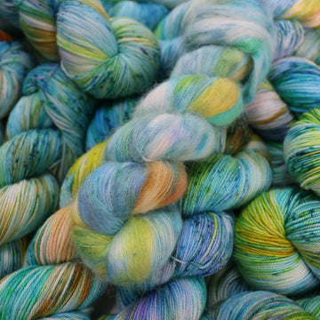 Suri yarn with light shades of blue and speckles of purple, with some light greens and browns. There are more skeins like this of different weights underneath.