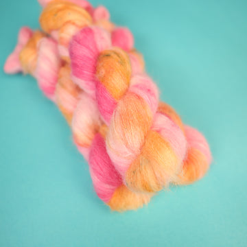 A couple of Suri Lace yarn skeins that are colored with a light pink, yellow, and orang e, on top of a light blue background.