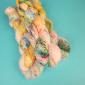 A light cream colored Suri yarn with different colored speckles all over and banana colored yellow at the top of the skein on top of a light blue background.