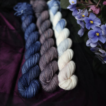 Three DK Mini skeins of yarn sitting besides each other. One on the left is colored a deep lake blue with the slightest undertones of mauve. The one in the middle is colored a plum grey with blue and dark grey color whispers. The final skein one the right is colored an elegant white with dramatic blue, the palest of mauve & amp grey color washes. All sitting on stop of dark blue dark purple and black fabric surrounded by colorful flowers.
