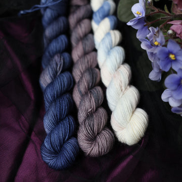 Three fingering mini skeins of yarn sitting besides each other. One on the left is colored a deep lake blue with the slightest undertones of mauve. The one in the middle is colored a plum grey with blue and dark grey color whispers. The final skein one the right is colored an elegant white with dramatic blue, the palest of mauve & amp grey color washes. All sitting on stop of dark purple and black fabric surrounded by colorful flowers.