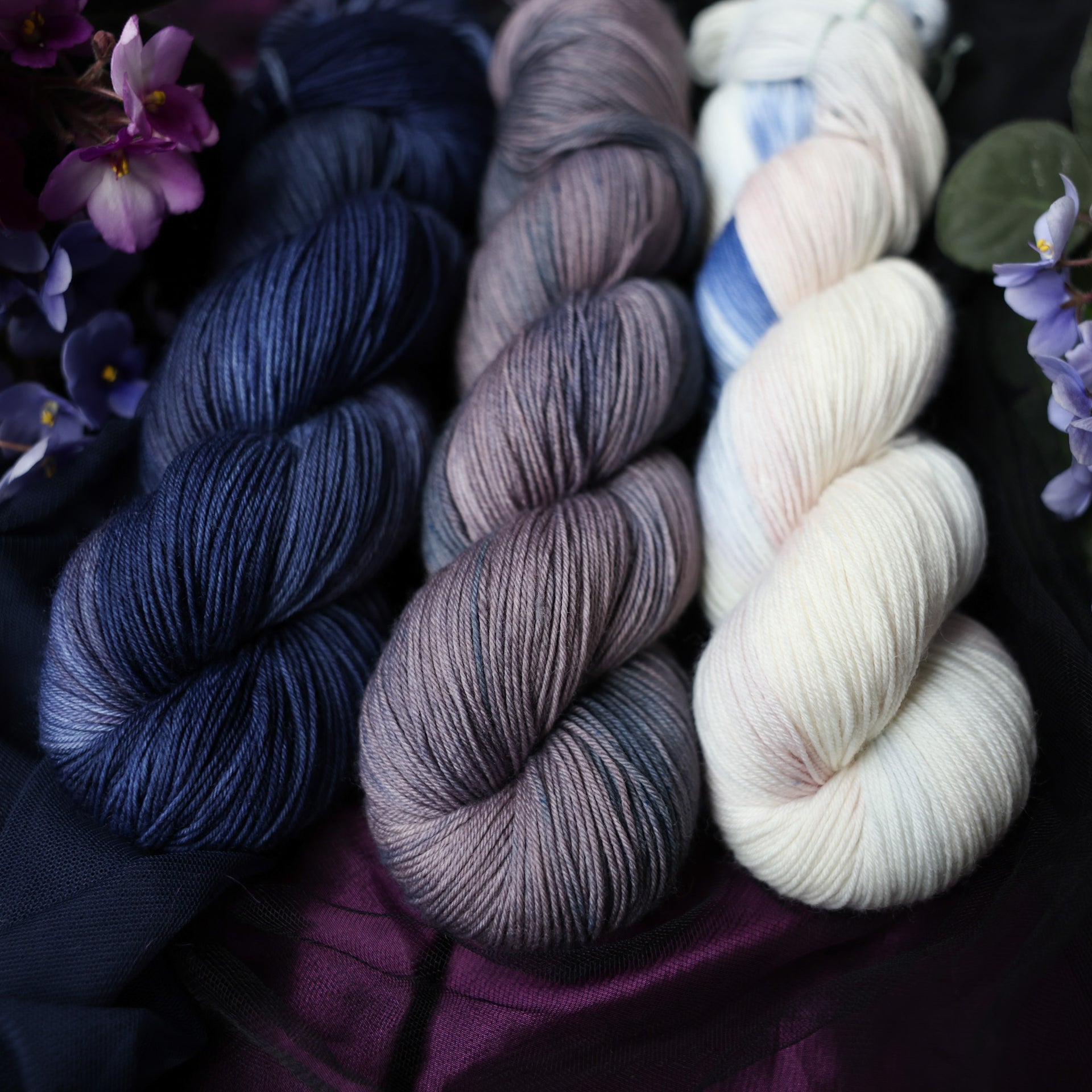 Three fingering skeins of yarn sitting besides each other. One on the left is colored a deep lake blue with the slightest undertones of mauve. The one in the middle is colored a plum grey with blue and dark grey color whispers. The final skein one the right is colored an elegant white with dramatic blue, the palest of mauve & amp grey color washes. All sitting on stop of dark blue dark purple and black fabric surrounded by colorful flowers.