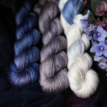Three Silk skeins of yarn sitting besides each other. One on the left is colored a deep lake blue with the slightest undertones of mauve. The one in the middle is colored a plum grey with blue and dark grey color whispers. The final skein one the right is colored an elegant white with dramatic blue, the palest of mauve & amp grey color washes. All sitting on stop of dark blue fabric surrounded by colorful flowers.