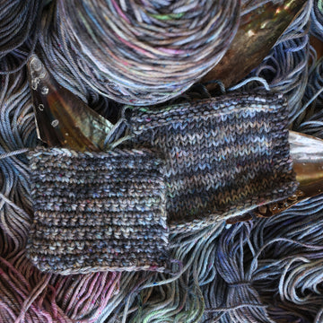 Crochet and knit swatches colored with different shades of grey and streaks of green, and dark pink, also there are also blue and purple speckles. These swatches are on top of unwound skeins of the same color way with some shells on top.