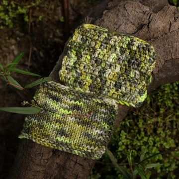 Knit and crochet yarn swatches colored with different shades of green and grey, on top of a tree limb that is hovering over a pond, with different mossy plants around the pond, and around the yarn.