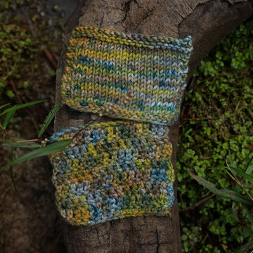 Knit and crochet swatches colored with green, yellow and grey colors, with blue speckles, on top of a tree limb that is hovering over a pond, with different mossy plants around the pond, and around the yarn.