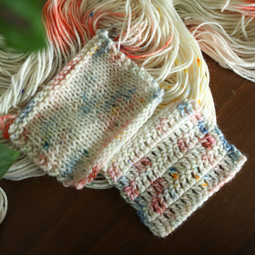 Crochet and Knit swatches of white yarn with mixed colors of light pink purple and red/orange and brown with speckles, on top of an unwound version of that same yarn. all on top of a dark wooden table with a couple plant leaves in the corner.
