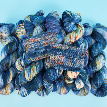 Different weights of a yarn skein colored with different shades of blue and white with multicolored speckles on the portion of the yarn that Is white. all on top of a light blue background and knit and crochet swatches of that yarn on top of the stack of yarn.