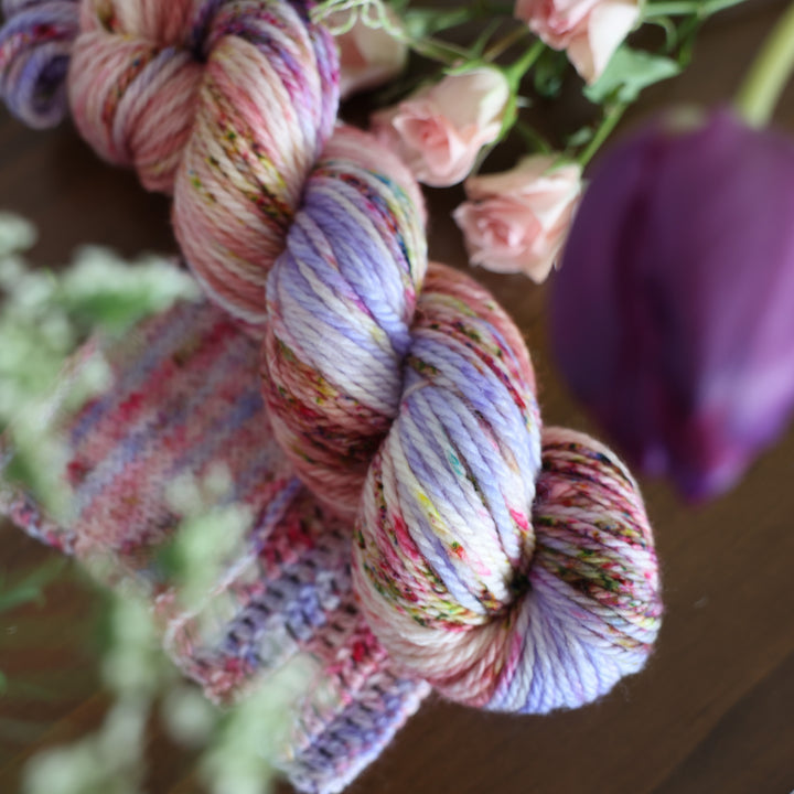 Bulky yarn and crochet and knit swatches colored different shades of light and dark pink with creme colors and a light lavender. This is on top of a brown table with some purple and light pink tulips around the yarn.